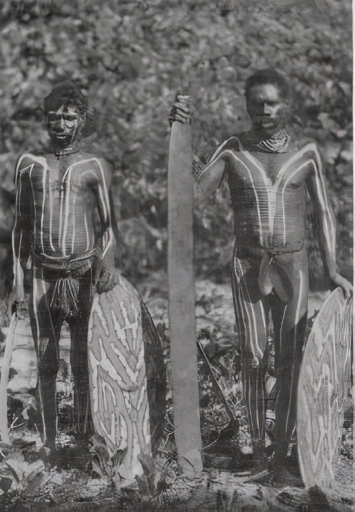 Two men with their weapons, a large battle axe, two painted shields, and a boomerang.