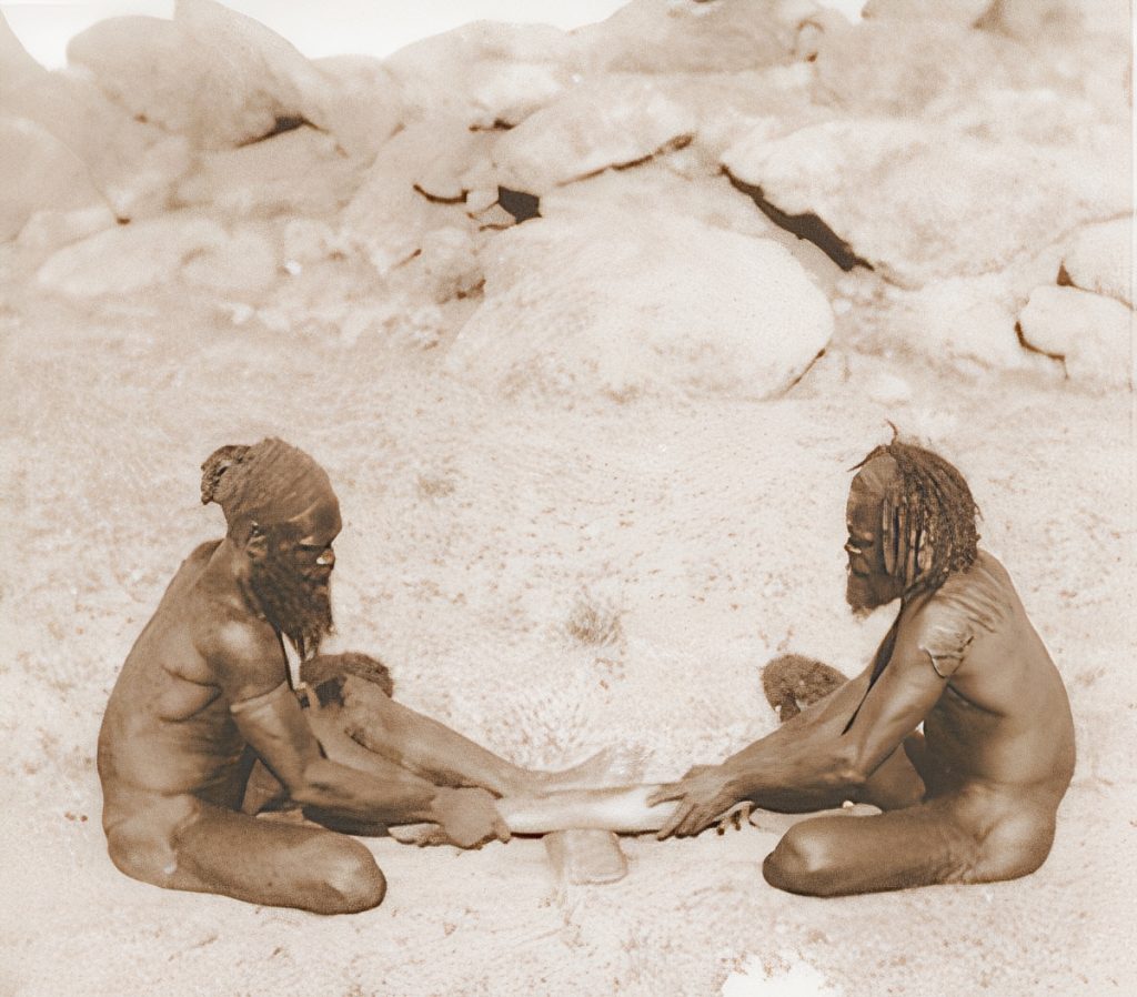 Two Aranda men in Central Australia rub the sharp edge of a hardwood spearthrower over a softwood shield to make an ember using the fire saw method.
